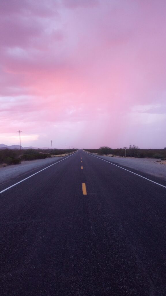 A 2-lane road vanishing into pink and grey clouds during late sunset. The desert of Baja California frames the road as it disappears to the horizon