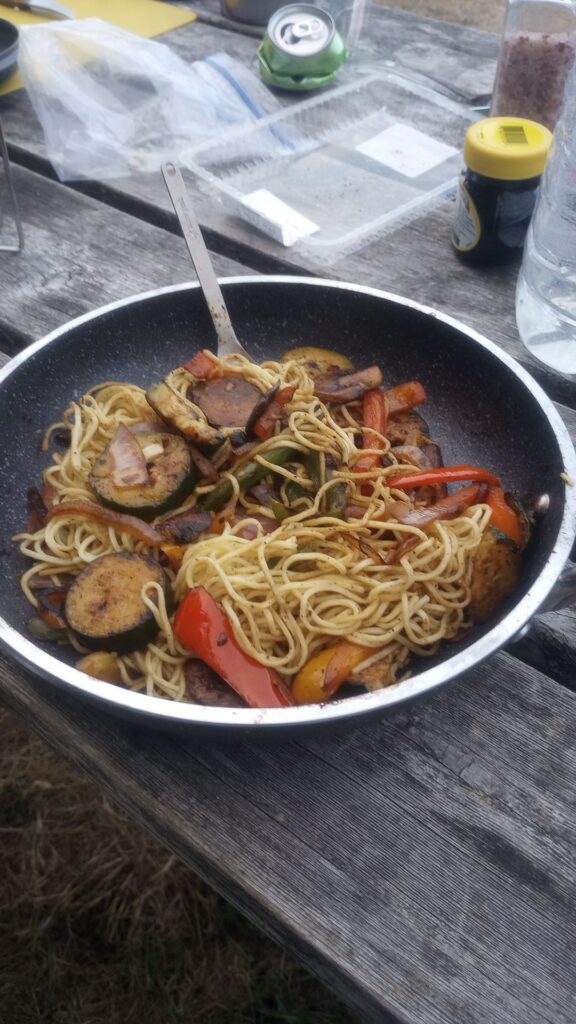 Camp fire fry up of noodles and vegetables sitting in a frying pan on a wooden table.
