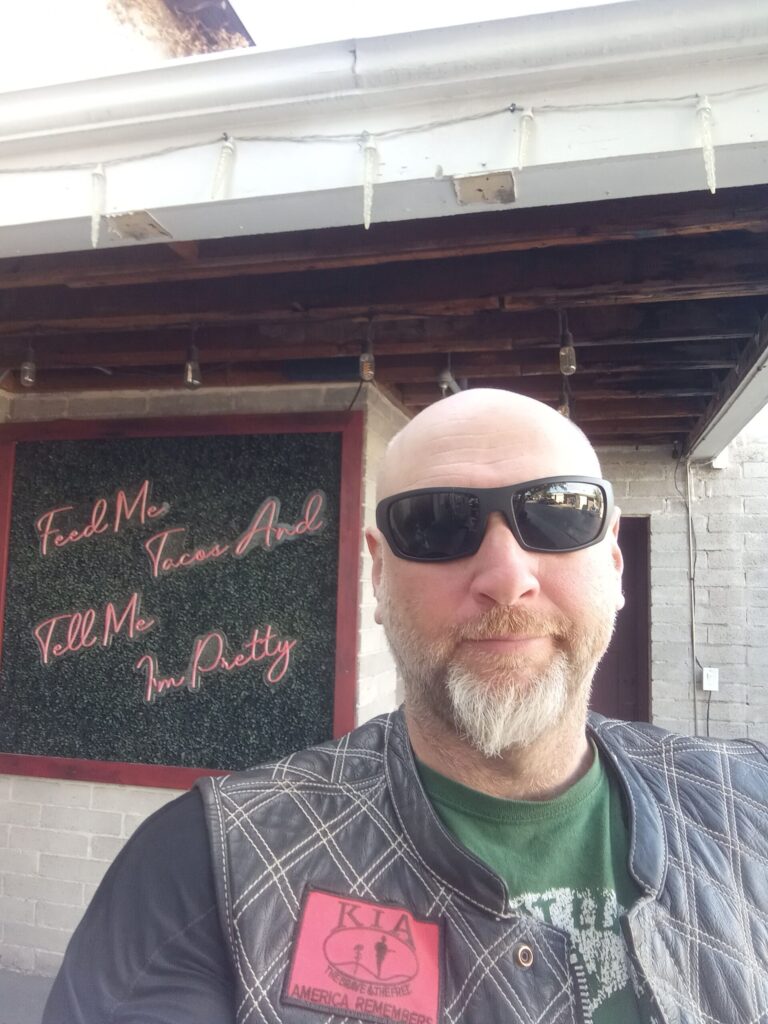 Selfie of a man in front of a sign reading "buy me tacos and tell me I'm pretty."