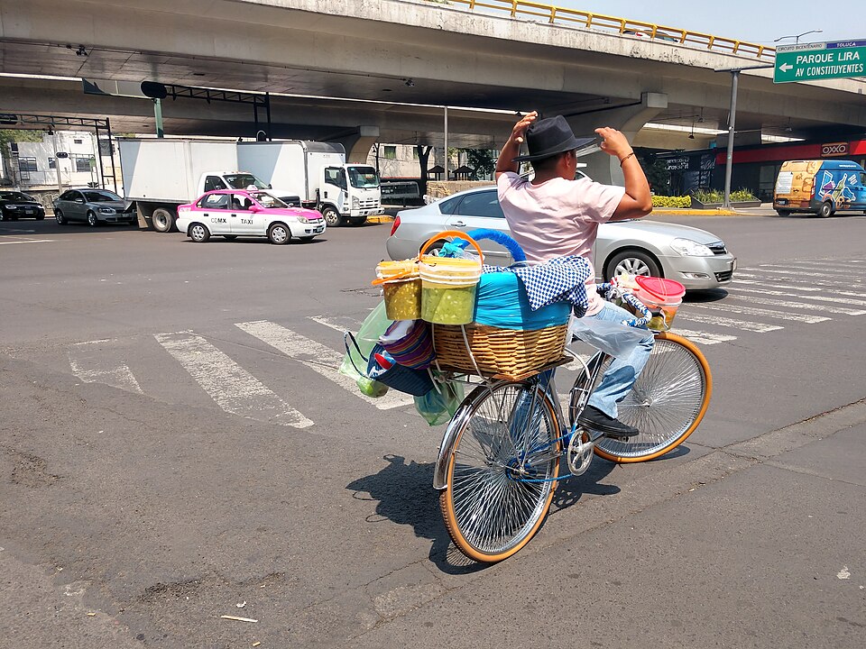 tacos de canasta vendor rides a bicycle on a busy street, with his basket of across on the back, along with jugs of salsa and bags for plates and napkins
