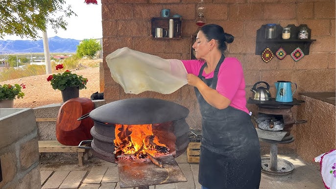 A woman on an outdoor patio works near an open flame with a convex curved steel top. She is working a massive flour tortilla in the air over the heat, arm outstretched, with the tortilla reaching from past her fingertips to mid-bicep