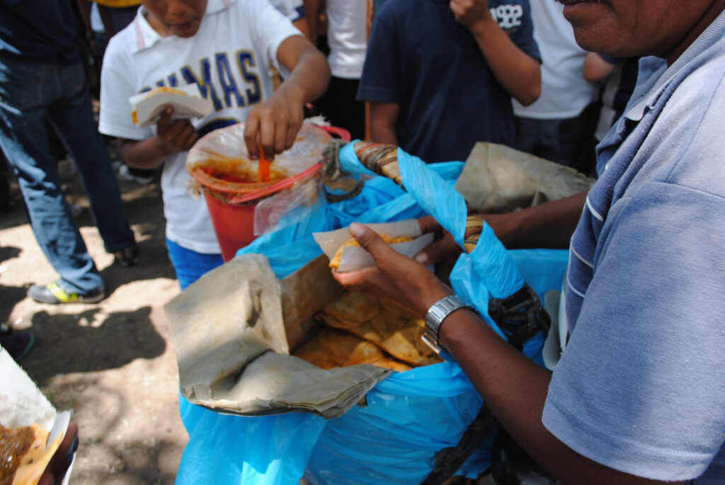 over the shoulder pic of a taquero selling tacos de canasta. The focus is on his hand, holding a taco, and behind is his open basket with its blue liner, and patrons scooping salsa from a lined pail.
