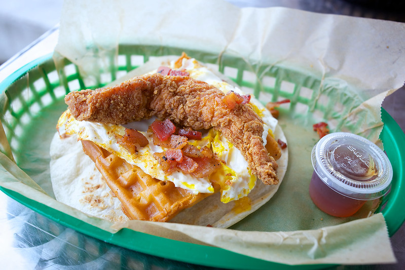 A taco in a basket with wax paper, but the filling is a chicken tender and half-wedge of waffle, with over-medium egg and crumbles of fresh bacon.