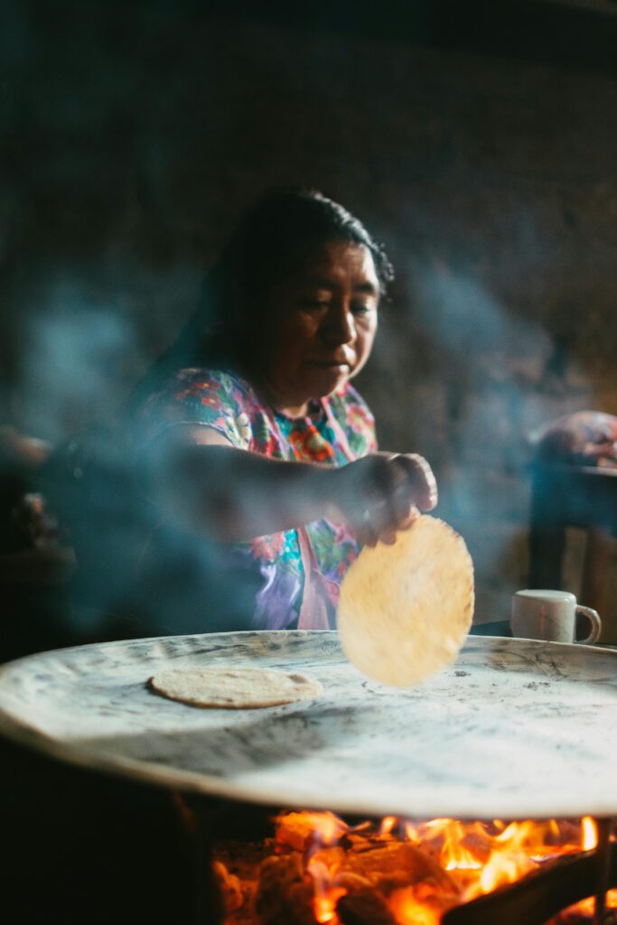 Mexican woman laying a tortilla onto a flat top piece of steel as smoke from an open fire beneath wafts up into frame. Late day light brings aliveness to a tranquil scene