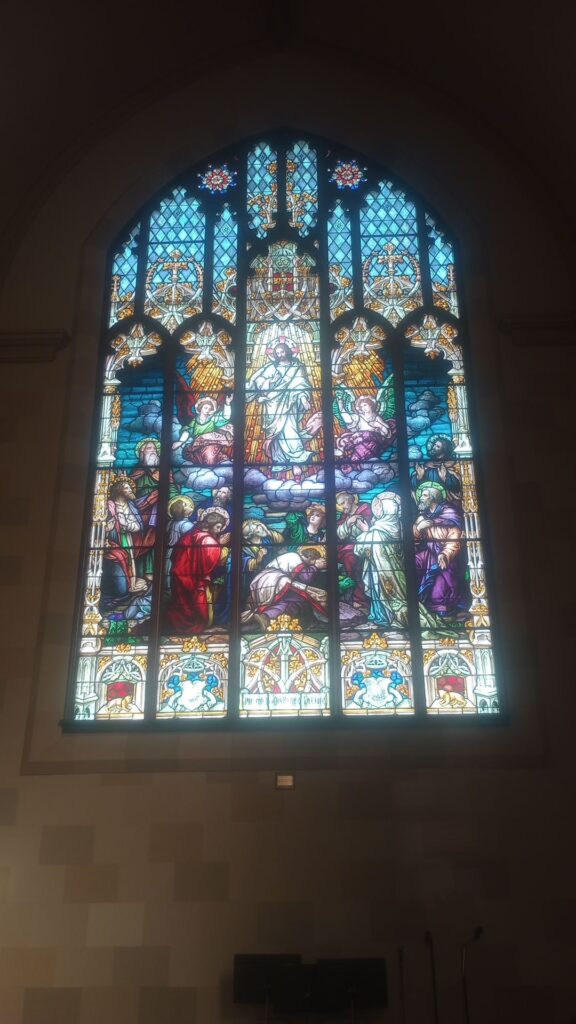 stained glass window depicting christ in the center with angels nearby and below a group that appear to be praying with head bowed or arms raised up toward him. Intricate patterns exist above and below the image, also part of the glass work